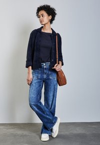 Marinefarbener, strukturierter Cardigan über einem dunklen Oberteil, kombiniert mit weiter geschnittenen blauen Jeans und weißen Sneakern. Eine braune Umhängetasche ruht auf einer Schulter.