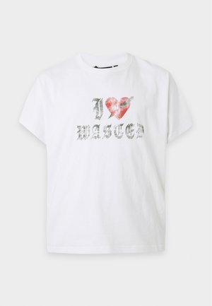 Млад мъж с къса коса, носещ бяла тениска с текст "I love Wasted" и черен панталон, изправен срещу обикновен светъл фон.