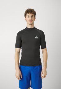 Czarny rash guard z krótkim rękawem, wykonany z elastycznego materiału, z małym szarym logo na piersi, w połączeniu z niebieskimi szortami kąpielowymi.