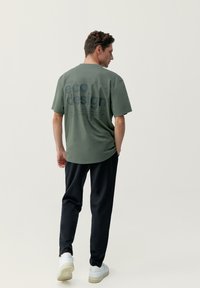 Zelené ekodesign tričko s veľkým čiernym textom na chrbte, kombinované s čiernymi nohavicami a bielymi teniskami. Jednoduchý, voľný strih.