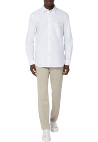 En man står klädd i en vit skjorta med knappar, beige byxor och vita sneakers, med händerna vilande vid sidorna, i en neutral miljö.