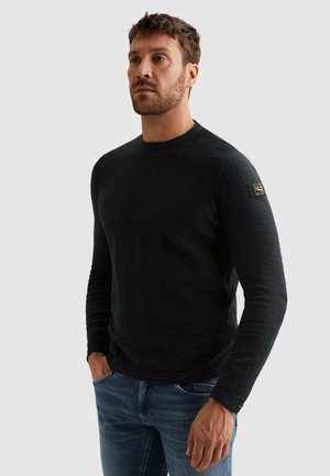 Moški v črnem puloverju z dolgimi rokavi z značko na zgornjem ročnem delu in modrih kavbojkah, stoji z rokami v žepih.