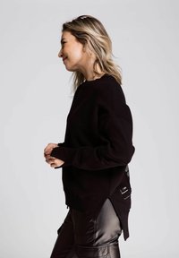 Schwarzer Sweatshirt mit lockerer Passform, gerippter Textur und seitlichen Schlitzen. Verfügt über ein einzigartiges Designelement auf der Rückseite. Kombiniert mit glänzenden, dunklen Hosen.