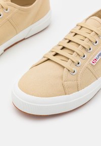 Beżowe płócienne trampki z teksturowaną powierzchnią, białą gumową podeszwą i srebrnymi oczkami. Posiadają logo marki po boku. Standardowy system sznurowania.