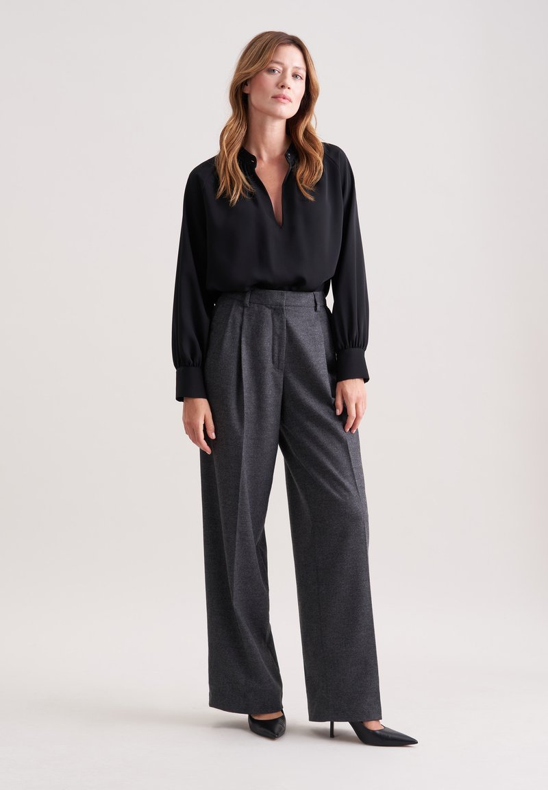 Blusa nera con scollo a V e maniche lunghe, abbinata a pantaloni grigi a vita alta e a gamba larga. Il modello indossa tacchi a punta neri.