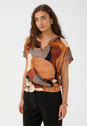 Une femme présente un chemisier à manches courtes aux motifs audacieux rouille, orange, beige et gris, associé à un pantalon noir, sur un fond uni.
