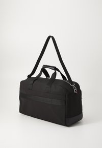 Sac de sport noir en tissu, doté de deux poignées supérieures, d'une bandoulière amovible et d'une bande de marque texturée à l'avant.
