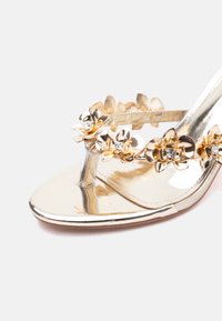 Χρυσό μεταλλικό σανδάλι με σχέδιο χιαστί λουριών, διακοσμημένο με χρυσά λουλούδια και διαφανείς στρας στο κέντρο των λουριών.