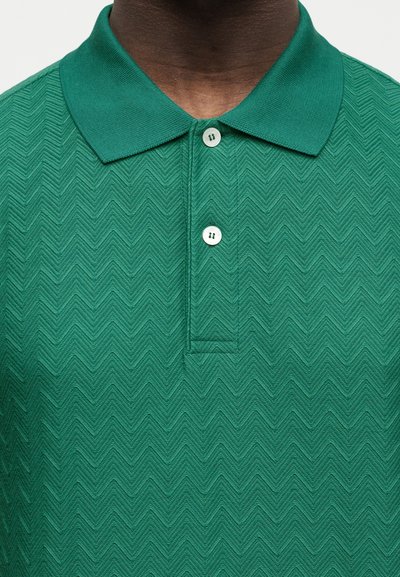 Άνδρας που φοράει πράσινο πικάζ με υφή και δύο λευκά κουμπιά κουμπωμένα στο γιακά.