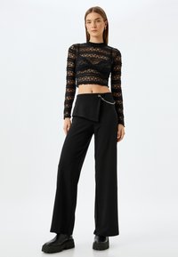 Zwarte kanten cropped top met lange mouwen en een geschulpte zoom, gecombineerd met hoge zwarte broeken met een zijklep en kettingdetail.