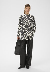 Must-valge abstraktse mustriga nööpidega särk, mida on kombineeritud kõrge vöökohaga mustadest nahkkulutükkidest pükstega ja musta nahast õlakotiga.