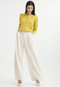 Geel, knopen-up gebreide trui met een kraag; witte, wijde broek; model draagt beige hakken; soepele texturen en een schoon ontwerp.