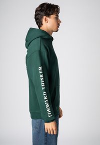 Grüner Kapuzenpullover mit Kängurutasche. Weißer Aufdruck "FORWARD THINKER" entlang des linken Ärmels. Weicher, strukturierter Stoff, lockere Passform.
