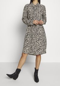 Robe à imprimé léopard en beige et noir, avec un devant à boutons, des manches longues et une coupe décontractée. Portée avec des bottes de cow-boy noires.