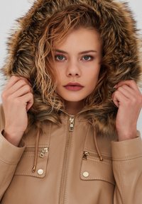 Tan faux leather jacket with a fur-lined hood, gold zippers, and pockets. Model holds the hood, showcasing the texture and detailing.