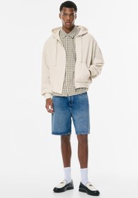 Beige Kapuzenpullover über einem grün karierten kurzärmeligen Hemd, kombiniert mit Jeansshorts und schwarzen Schlüpfschuhen mit weißen Sohlen.