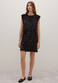 Jeune femme portant une robe mini sans manches noire ornée de sequins et des chaussures plates noires brodées, se tenant devant un fond uni clair.