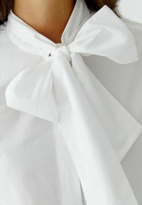 Witte blouse met een grote strik kraag van gladde, stevige stof. De textuur is zacht en het ontwerp heeft een schone, minimalistische esthetiek.