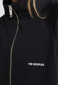 Černá bunda na zip s vysokým límcem, stříbrným zipem a bílým vyšitým textem "THE KOOPLES" na přední straně. Hladká textilie.