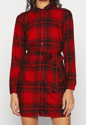 Femme portant une robe chemise à carreaux rouge et noir avec manches longues et ceinture nouée à la taille.