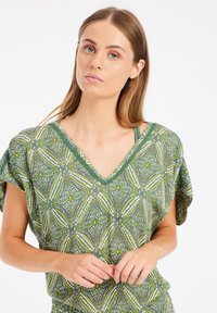 Zelená blúzka s geometrickým vzorom a V-výstrihom, krátkymi rukávmi, s žltými a bielymi akcentmi. Ľahký textil.