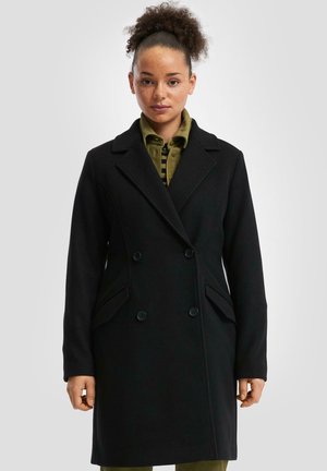 Manteau noir à double boutonnage avec des revers crantés, poches latérales et une texture lisse, porté par-dessus une chemise olive.