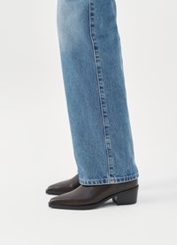 Stivali alla caviglia in pelle marrone con punta a punta e tacco a scatola, abbinati a jeans dritti blu chiaro, che mostrano un design pulito e semplice.