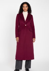 Bordeauxroter langer Mantel aus strukturiertem Stoff, mit einem einzelnen Knopf, Revers mit Kerbkragen und zwei Vorderentaschen. Kombiniert mit grauen Hosen.