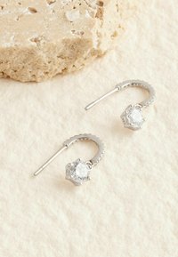 Silverfärgade ringörhängen med hexagonala klara stenar, med en texturerad design och slät metallyta, mot en neutral bakgrund.