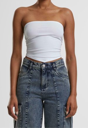 Osoba nosící bílý top bez ramínek a modré volné džíny s vysokým pasem a nákladními kapsami, stojící před jednobarevným pozadím.