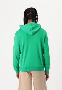 Felpa verde realizzata in tessuto morbido, con cappuccio, maniche lunghe e vestibilità rilassata. Lo sfondo è bianco semplice. I pantaloni corti sono beige.