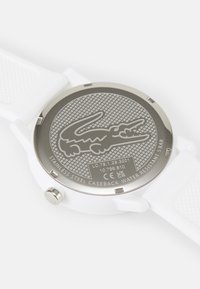 Reloj con correa de silicona blanca y fondo de acero inoxidable, que presenta una superficie texturizada y un logo de cocodrilo grabado. Resistente al agua hasta 5 bares.