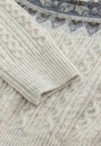 Világos bézs gyapjúpulóver, amely kábelmintás textúrával és szürke-barnás mintázatú vállrésszel rendelkezik. Rugalmas mandzsettákkal és aljjal készült.