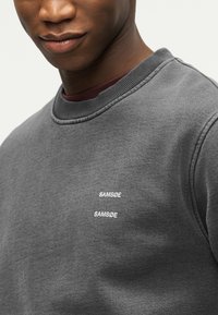 Κοντινό πλάνο ενός ατόμου που φοράει γκρι φούτερ με το κείμενο "SAMSØE" τυπωμένο δύο φορές στο στήθος με λευκά γράμματα.