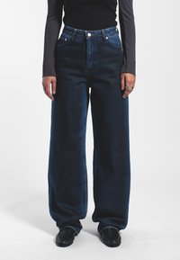 Jeans de pernera ancha en azul oscuro que presentan una cintura alta, cierre de botón y un diseño de cinco bolsillos, elaborados con tela vaquera de textura suave.