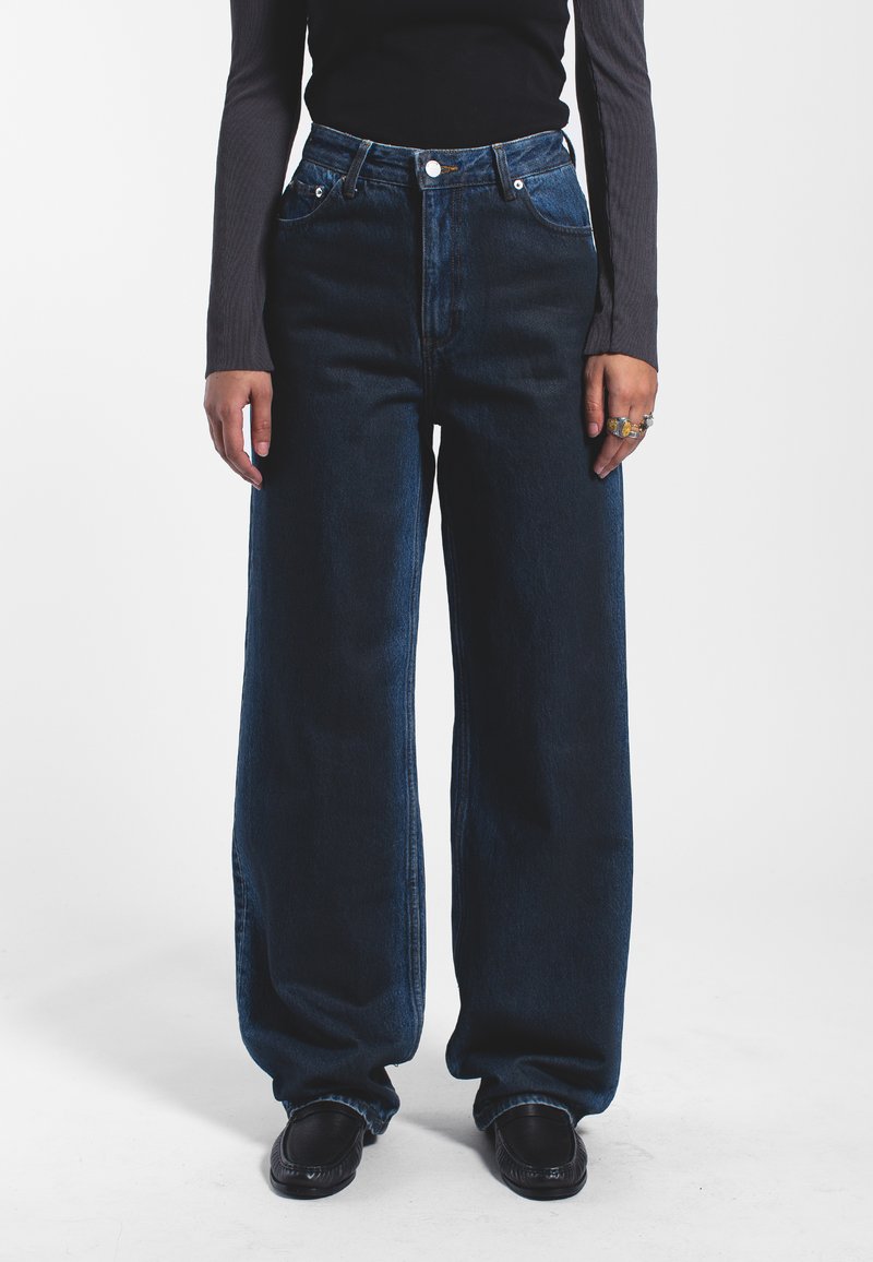 Jeans de pernera ancha en azul oscuro que presentan una cintura alta, cierre de botón y un diseño de cinco bolsillos, elaborados con tela vaquera de textura suave.