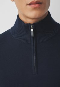 Marineblauer Reißverschluss-Pullover mit geripptem Kragen, strukturiertem Stoff und silbernem Reißverschluss. Nahaufnahme, die den Kragen und die obere Brust hervorhebt.