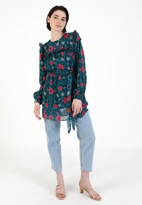 Koszulka w kwiatowy wzór w kolorze teal z czerwonymi i różowymi kwiatami, z falbanami na ramionach, związanym w pasie i długimi rękawami; zestawiona z jasnymi dżinsami.