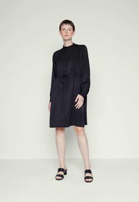 Μαύρο φόρεμα μέχρι το γόνατο με μακριά μανίκια, συγκεντρωμένη λαιμόκοψη, ελαστική μέση και λεπτομέρεια με κορδόνι. Λείος υφάσματος με υποτονική γυαλάδα.