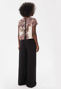 De ingekorte blouse heeft een paisleypatroon in roomwit, bordeauxrood en blauw, met korte mouwen en een knoopsluiting aan de achterkant. Gekocht in combinatie met zwarte wijde broek.