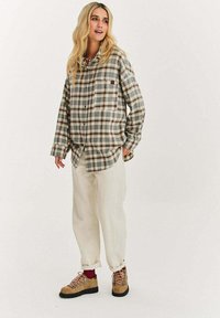 Kobieta z długimi blond włosami, ubrana w oversize'ową beżową koszulę w kratę, luźne kremowe spodnie, czerwone skarpetki i beżowe buty trekkingowe, stojąca na białym tle.