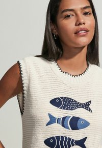 Vit stickad ärmlös topp med tre blå fiskmotiv i olika mönster. Urringningen har ribbat kant med svarta detaljer.