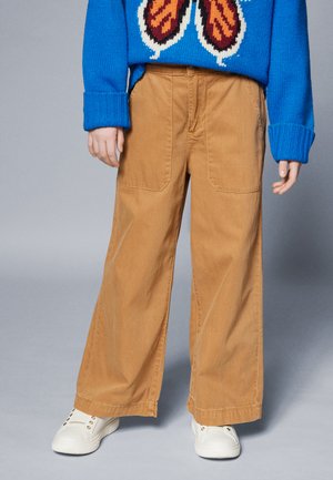 Brede beige katoenbroek met twee voorzakken, gecombineerd met een blauwe trui met een vlinderontwerp en witte sneakers.