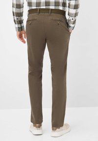 Pantaloni chino marroni con una texture liscia, dotati di due tasche posteriori con dettagli in bottone. Indossati con una camicia a quadri e scarpe casual.