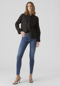 Svart knäppt skjorta med två bröstfickor, kombinerad med blå skinny jeans. Modell som bär klara högklackade sandaler. Enkel bakgrund.