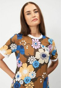 Kukallinen jersey-mekko, jossa on ruskea tausta, sinisiä, vaaleanpunaisia ja valkoisia kukkia, sekä valkoinen ribattu kaula-aukko ja hihat.