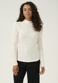 Langärmliges Oberteil aus strukturiertem, elfenbeinfarbenem Stoff mit hohem Kragen und floralem Muster. Kombiniert mit schwarzen Hosen, was einen kontrastierenden Look ergibt.