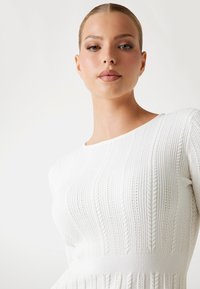 Pull en tricot blanc avec une texture côtelée, doté d'un col rond et de motifs en torsades verticaux, mettant en avant une silhouette ajustée.