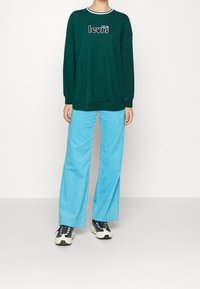 Felpa verde con logo Levi’s bianco e nero, abbinata a pantaloni larghi in velluto a coste azzurro chiaro e sneakers bianche e nere.