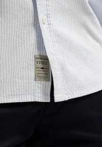 Jasnoniebieska koszula w paski z białymi guzikami i metką marki, wykonana z tkaniny o teksturowanym wykończeniu, zestawiona z czarnymi spodniami.